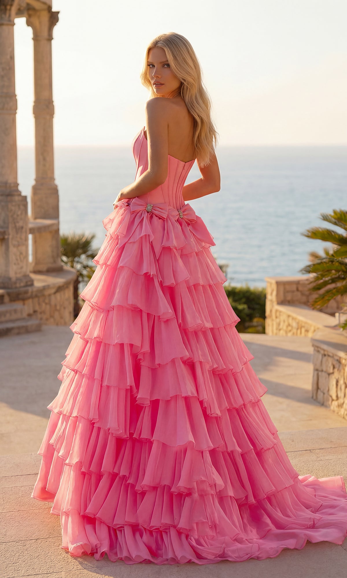 A woman stands outdoors by the sea, her back partially turned, wearing the PromGirl 250071 Tiered Prom Ball Gown with bows—a strapless, floor-length chiffon dress.